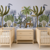 Neutral jungle nursery mural wallpaper featuring illustrated palm trees, tropical leaves, and exotic animals including zebra, antelope, monkey, parrots, toucans, and elephants. Styled in a twin nursery with two wooden cribs and neutral changing unit.