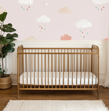 A nursery room with the Pink Clouds Mural on the wall, featuring a gold crib in the foreground and a plant to the left.